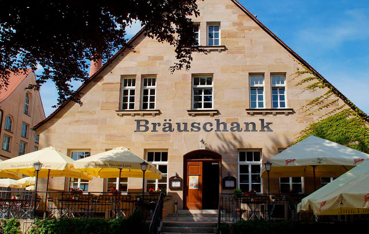 Ein sonniger Tag an einem sandsteinernen Gebäude mit der Aufschrift Bräuschank mit Sonnenschirmen davor.