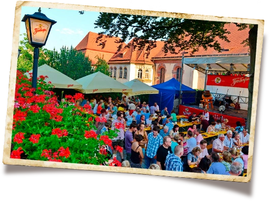 Viele Menschen sitzen draußen zusammen an Tischen und genießen ein Straßenfest. Rote Blumen und ein historisches Gebäude sind sichtbar.