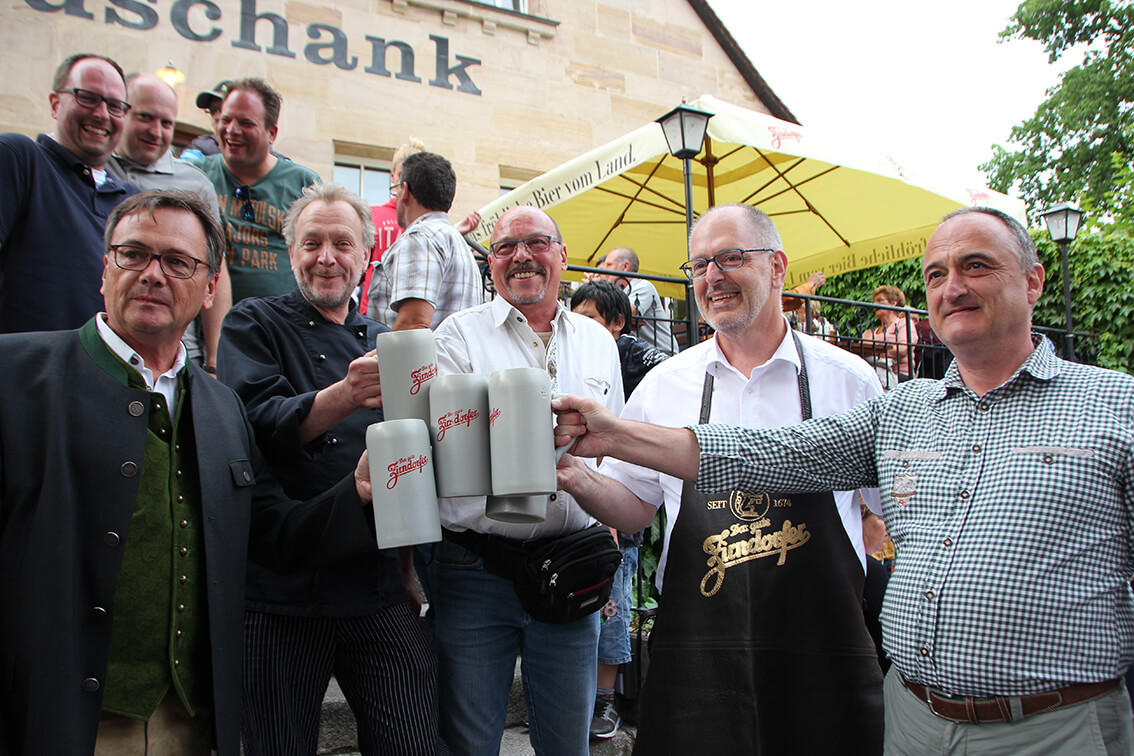 Mehrere Männer stehen lächelnd vor einem Restaurant und stoßen mit Bierkrügen an.