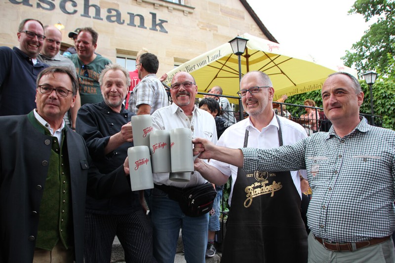 Mehrere Männer stehen lächelnd vor einem Restaurant und stoßen mit Bierkrügen an.