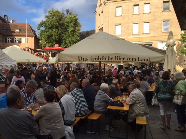 Menschen sitzen draußen an Tischen unter Sonnenschirmen und genießen Speisen und Getränke.