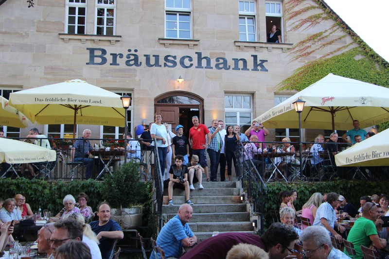 Menschen sitzen und stehen vor einem hellen Gebäude mit Sonnenschirmen.