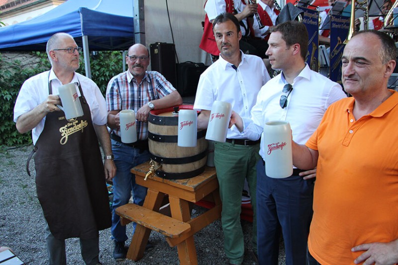 Fünf Männer stehen um ein Holzfaß, halten große Bierkrüge und lächeln.